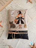 Finished cross stitch pillow of Moonlight Mischief featuring a smiling little witch holding a jack-o'-lantern bucket beside a gray cat under a crescent moon. Halloween cross stitch pattern by Luminous Fiber Arts.