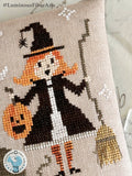 Close-up of the Moonlight Mischief Halloween cross stitch pattern featuring a smiling little witch with orange hair, a jack-o'-lantern bucket, and a rustic broom stitched on linen.